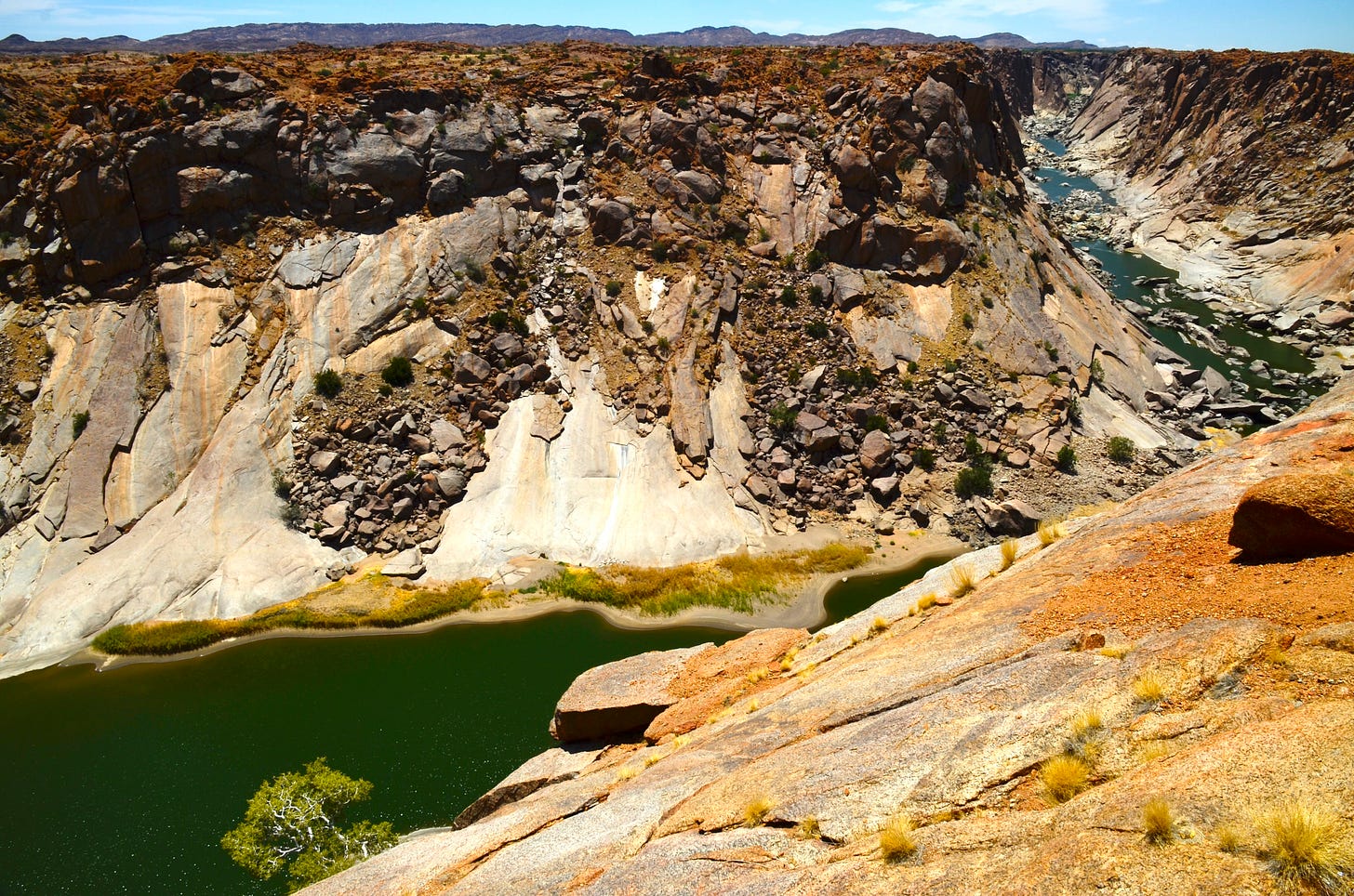 The photo showcases a breathtaking landscape of a deep canyon in Augrabies Falls National Park, South Africa. Steep, rugged cliffs composed of layers of brown, orange, and grey rock tower above a winding, greenish river that cuts through the canyon floor. The rocky walls are scattered with boulders and patches of sparse vegetation, highlighting the arid, harsh environment. The water below appears calm, with a small, verdant oasis of green shrubs and a lone tree thriving against all odds at the edge of the river, symbolizing resilience amidst the rugged terrain. The view is both awe-inspiring and humbling, evoking a sense of timelessness and natural beauty. The photo showcases a breathtaking landscape of a deep canyon in Augrabies Falls National Park, South Africa. Steep, rugged cliffs composed of layers of brown, orange, and grey rock tower above a winding, greenish river that cuts through the canyon floor. The rocky walls are scattered with boulders and patches of sparse vegetation, highlighting the arid, harsh environment. The water below appears calm, with a small, verdant oasis of green shrubs and a lone tree thriving against all odds at the edge of the river, symbolizing resilience amidst the rugged terrain. The view is both awe-inspiring and humbling, evoking a sense of timelessness and natural beauty.