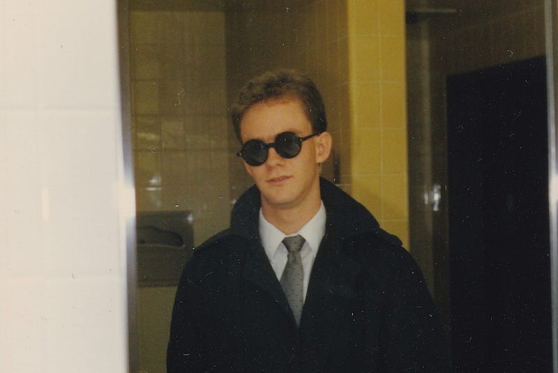 A young man in a black trenchcoat and dark sunglasses poses in front a mirror in a restroom.