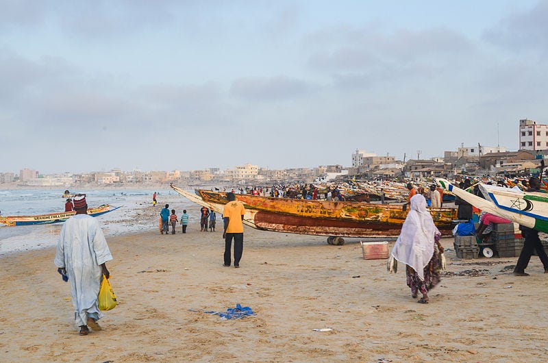 A picture of a crowded beach in Senegal, where surf and sand meet fishing boats and a distant skyline.