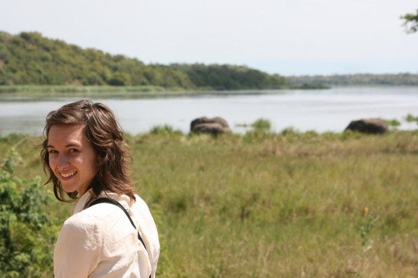 Kaitlin standing near water in Uganda, smiling, her hair at her shoulders, wearing a cream shirt. She is about 20 years old 