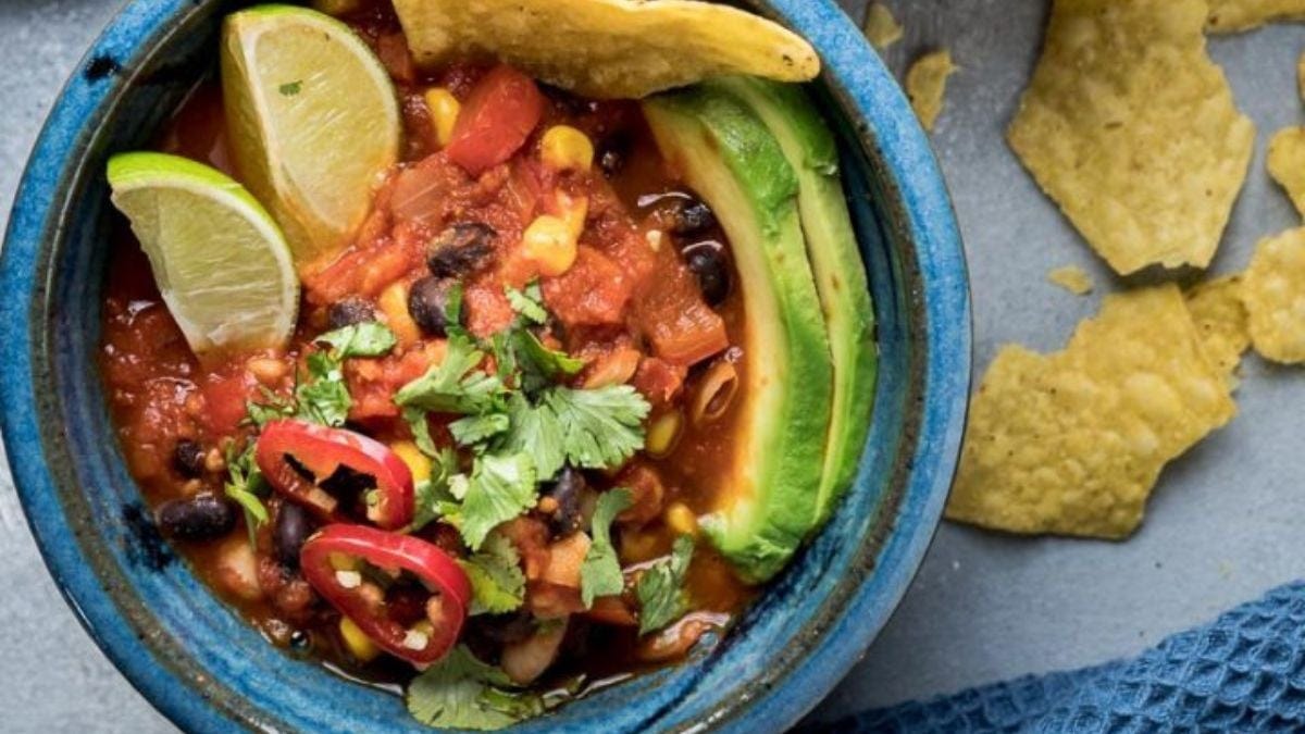 A vibrant bowl of mexican-inspired soup with black beans, tomatoes, avocado slices, and cilantro, garnished with lime wedges and red chili slices, served with broken tortilla chips on the side.
