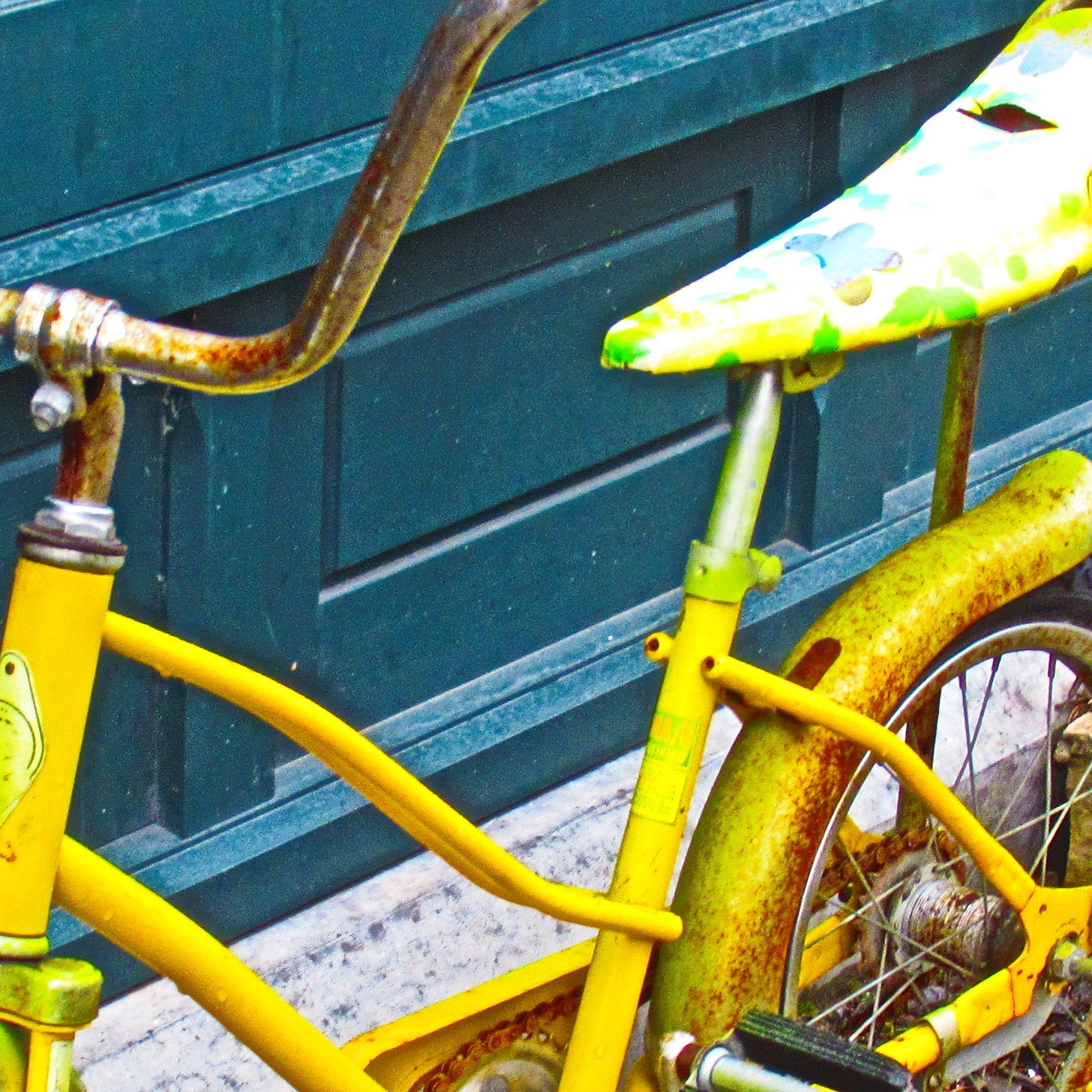 A rusting yellow bike with a bannana seat with a slit in it next to a teal blue facade for a shop.
