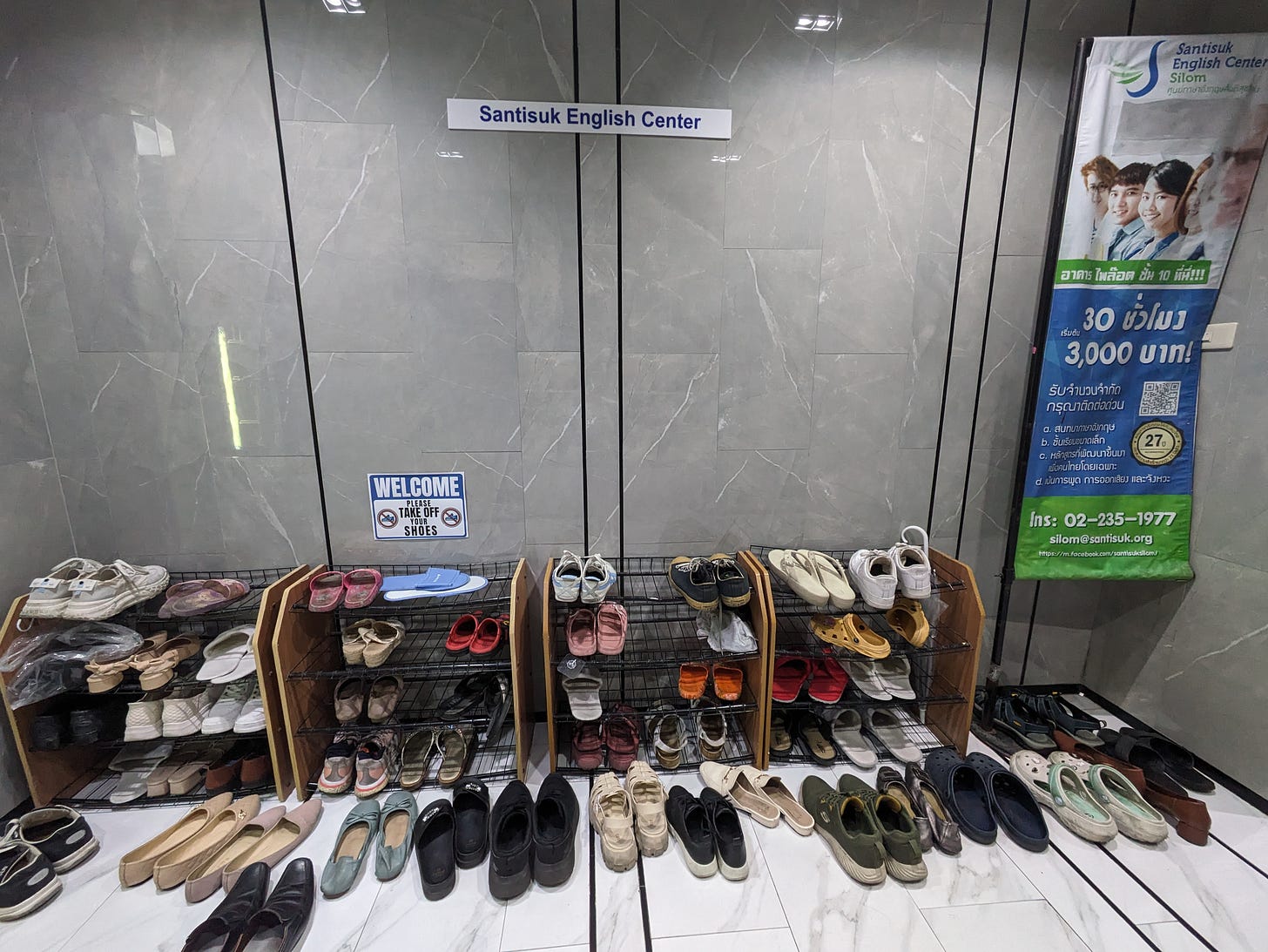 A large gathering of shelves and slippers on a white marble floor, set up against a gray marble wall labeled with Santisuk English Center