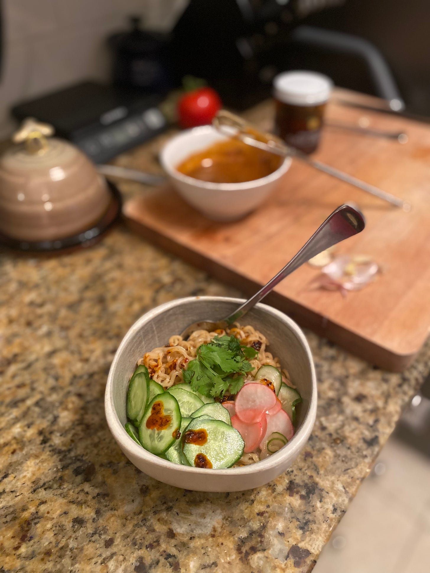 A bowl of instant noodles in sesame sauce, with cucumbers, pickled radish, cilantro, and chili crisp on top. In the background is the cutting board, and on it is a white bowl with the rest of the sauce and a whisk, a pile of garlic skins, and a jar of chili crisp.
