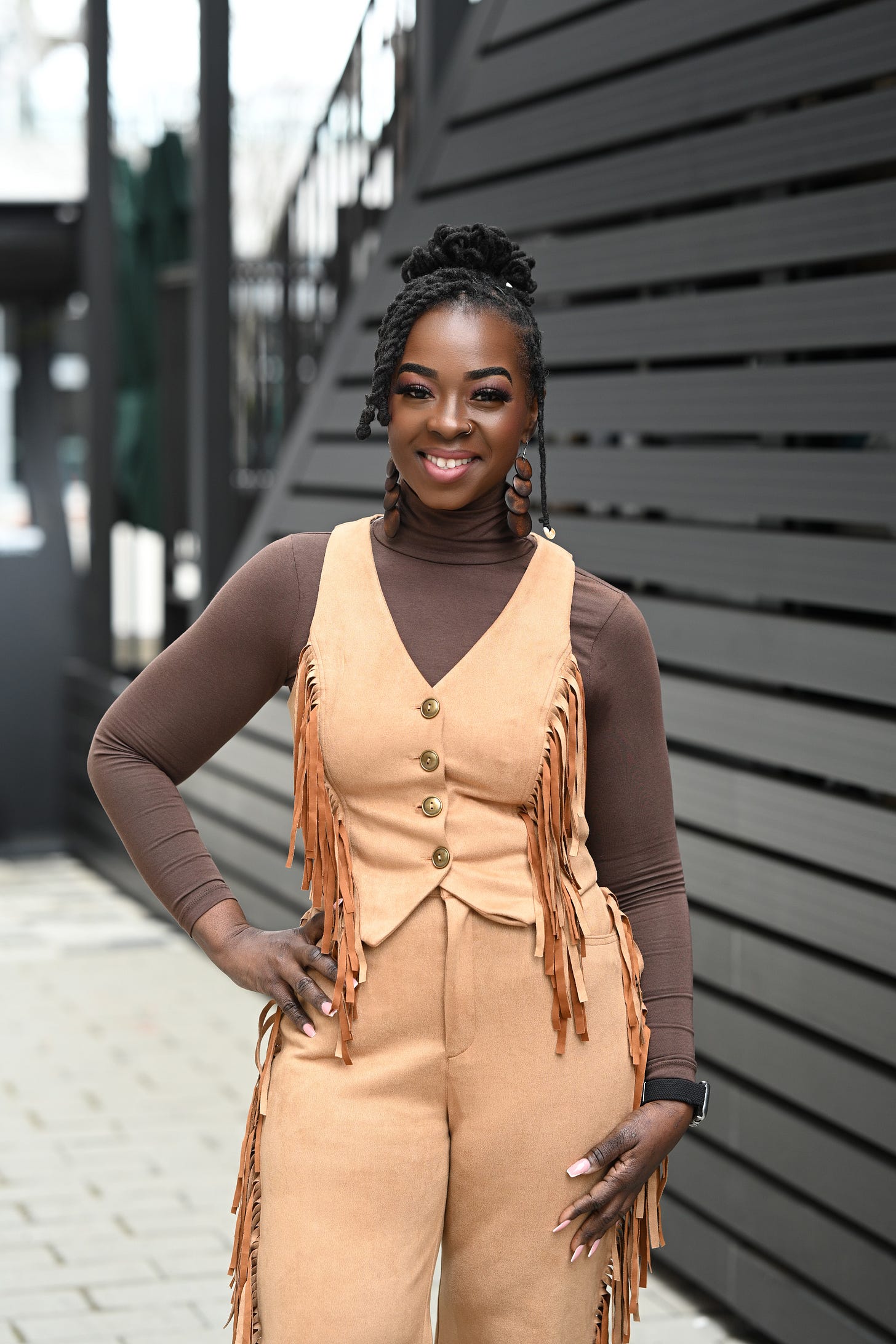 Monique McFadden, a chef, stands outdoors with a radiant smile. She wears a brown turtleneck and a tan suede vest with fringe details, paired with matching high-waisted pants. Her twisted locks are styled in an updo, and she accessorizes with bold brown earrings and a smartwatch. The background features modern black paneling and an urban walkway. Photo by Nakeshia Shannon.