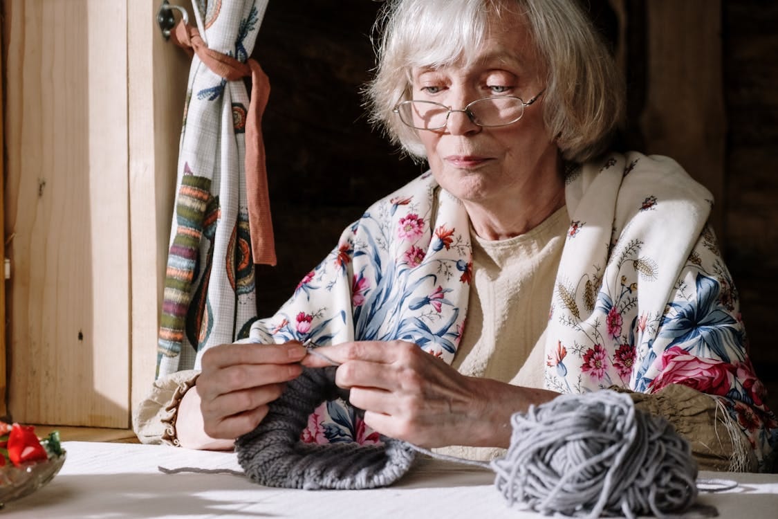 Free Senior woman knitting with gray yarn near a sunlit window, showcasing peaceful retirement activities. Stock Photo