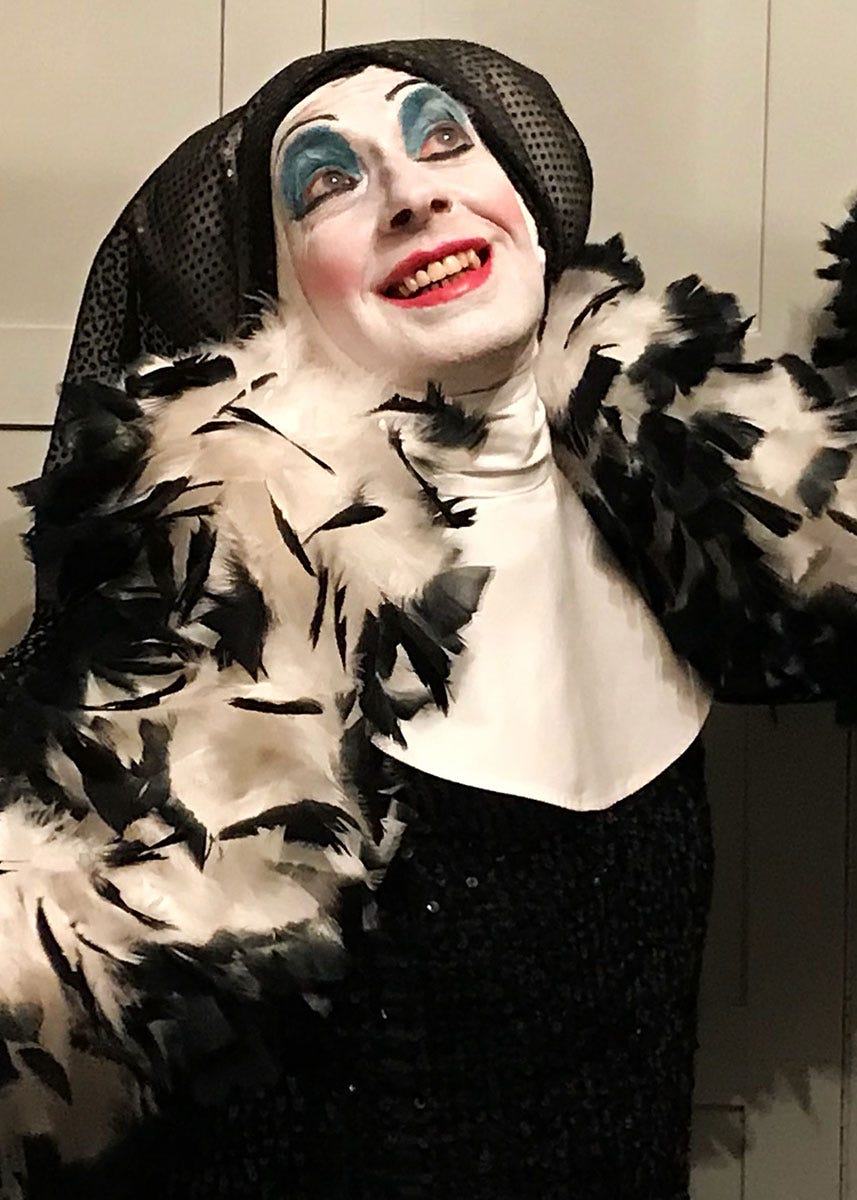 A person in sequined black dress, white and black boa, black nun's habit, and white face paint with exaggerated blue eye shadow and pink blush, smiling.