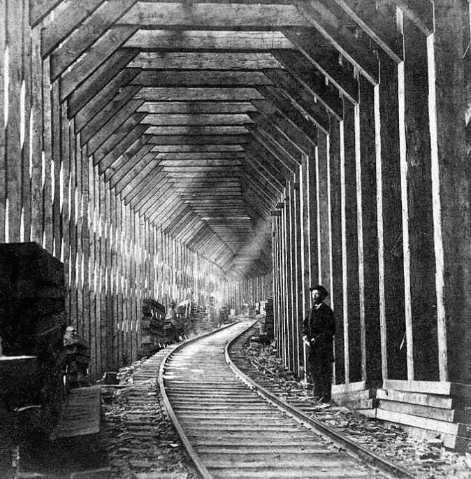 Old black and white photo of the railway sheds over Donner Pass. The sheds are tall, rectangular structures constructed of heavy wood rectangular posts that follow the curve of the railroad in the center of the frame. The posts are joined at the top by a beam and braced by triangular pieces. Sunlight is entering the sheds from the left and top right, suggesting that they are not completely sealed. A man stands beside the tracks in the foreground. And unused wooden beams and posts are stacked up along the shed, right up against the standing posts so as not to impede trains passing through.