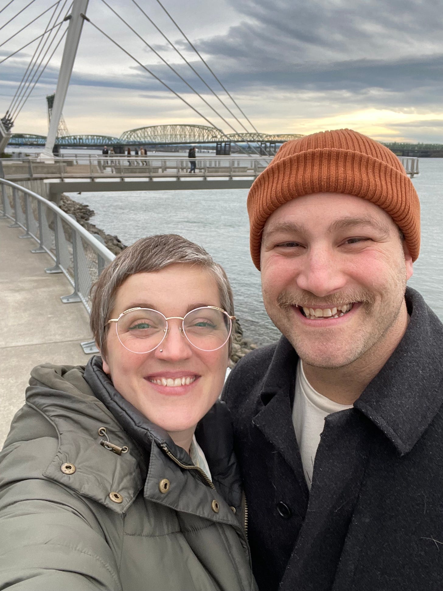 a white couple in their late 30s is standing in front of a river. The man has a mustache and is wearing a red beanie and is smiling. The other person has short graying hair and glasses and is also smiling. they look very happy together, because they are