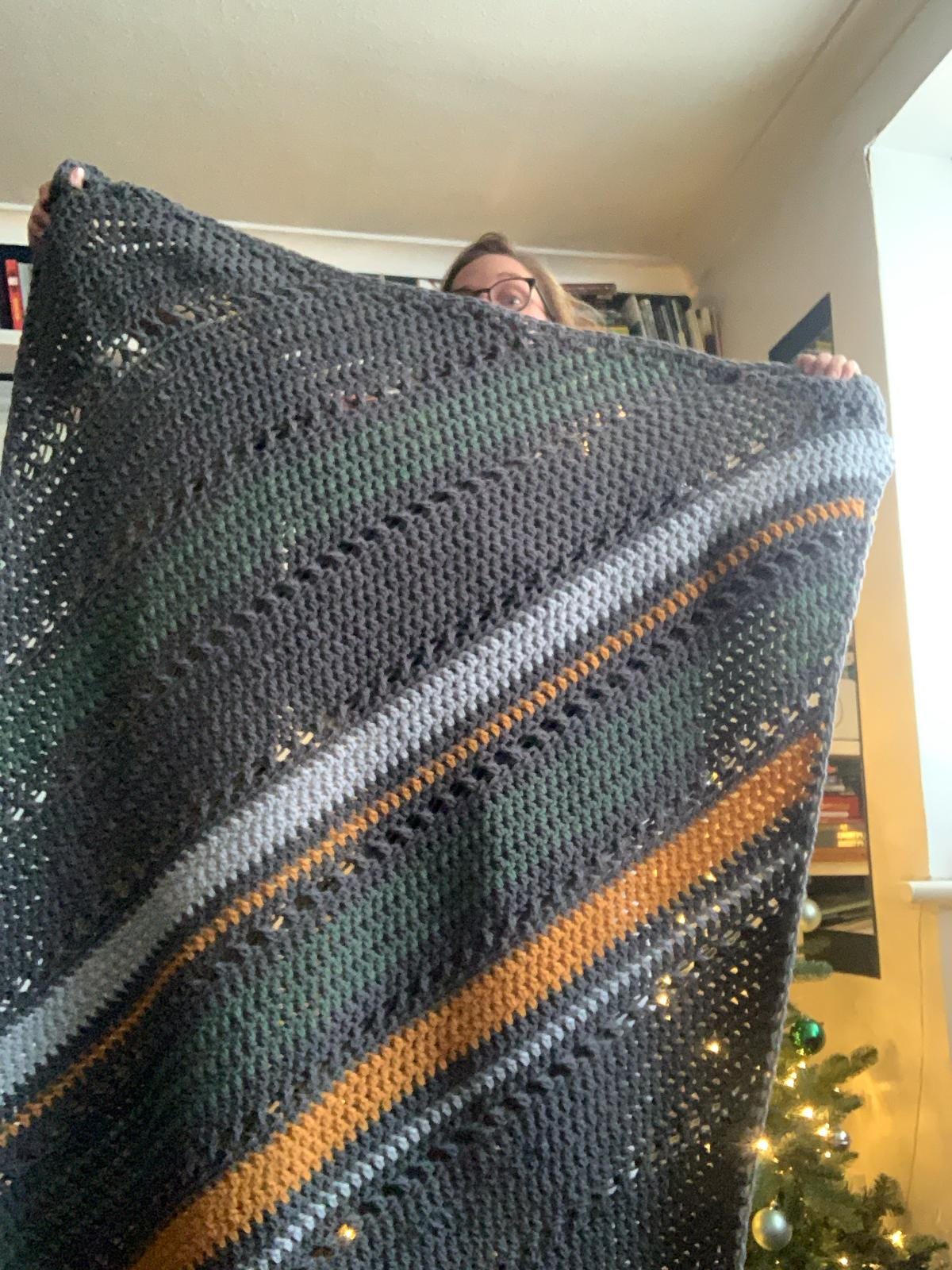 Jo holds up a grey, green and yellow striped crochet blanket as tall as her, standing in front of a Christmas tree.