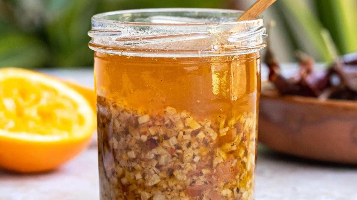 A glass jar filled with homemade orange marmalade featuring visible bits of orange zest, with a wooden spoon inside. half an orange and a bowl of dates are in the background on a marble surface.