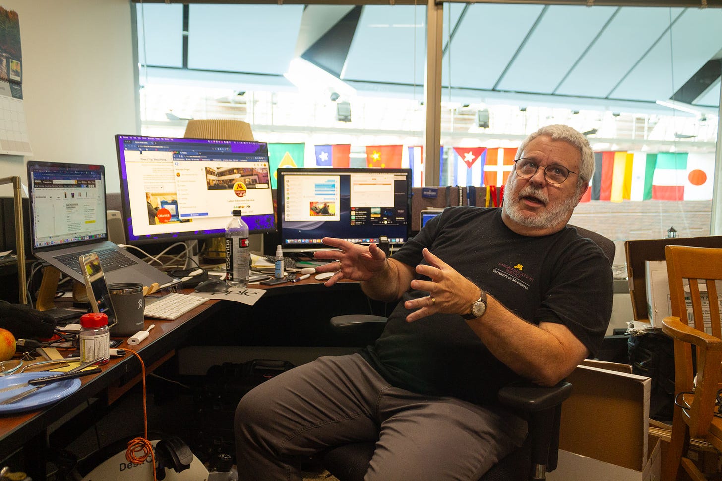 a white man with white hair and scruff wearing glasses and a black tee shirt sits at a desk with three monitors with his hands gesturing to the left of the image