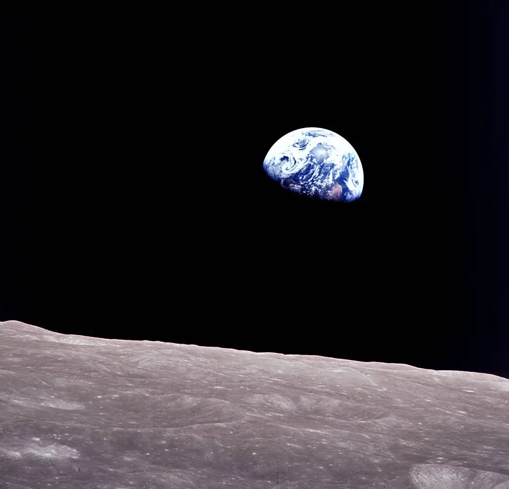 A view of the Earth against the blackness of deep space. The planet is an orb of swirling white and blue, its bottom half obscured by the darkness of the Moon's shadow. In the foreground is the rocky grey terrain of the Moon. 