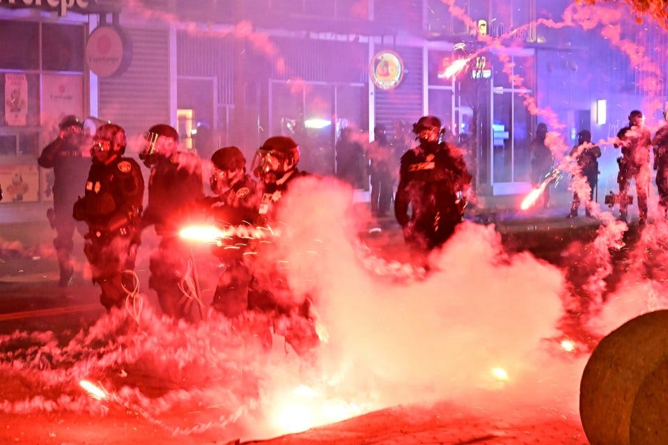 Police officers in riot gear during a protest amidst fireworks and smoke.