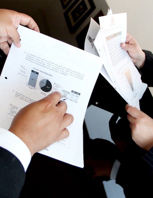 Two sets of hands going over a report. One is pointing at a graph and wearing a business suit, while the other set of hands is reading over the rest of the report.