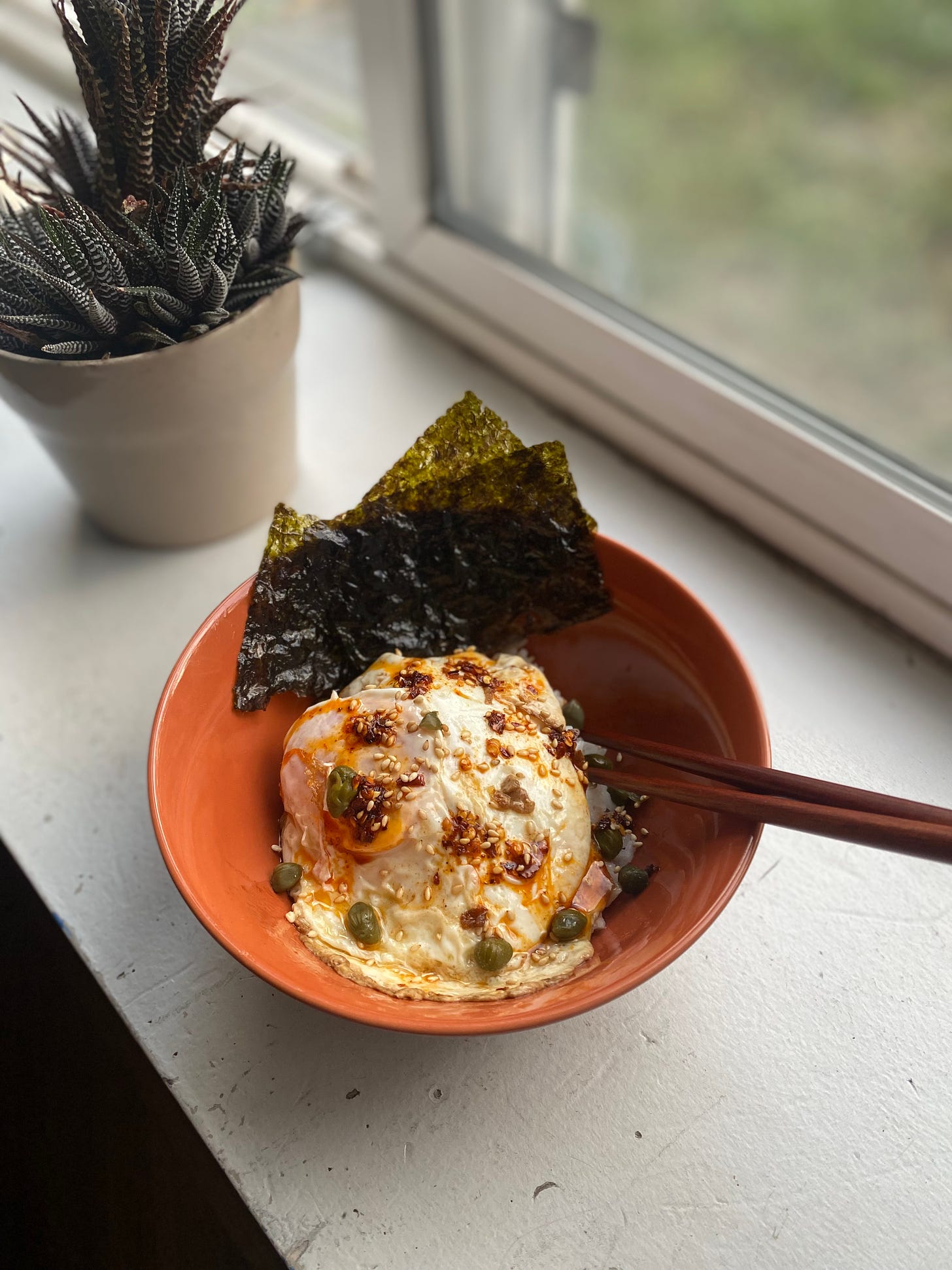 An orange bowl with an over easy egg on rice, topped with capers, chili oil, and sesame seeds. Several pieces of gim are at the back edge of the bowl, and chopsticks are sticking out of the side. The bowl is on a windowsill in front of a plant.