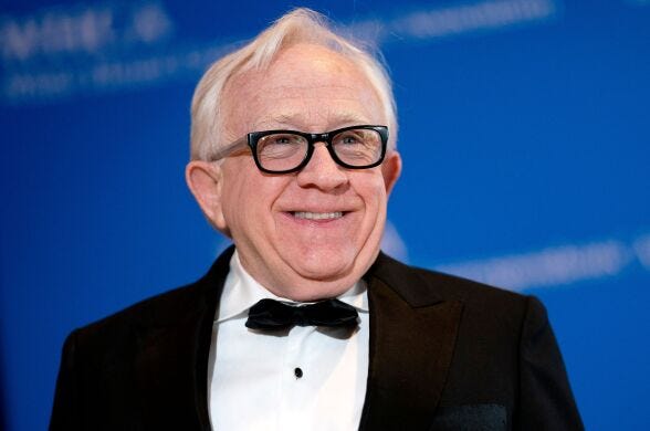 A man with light-tone skin has silver hair and wears dark rim glasses and a classic tux.