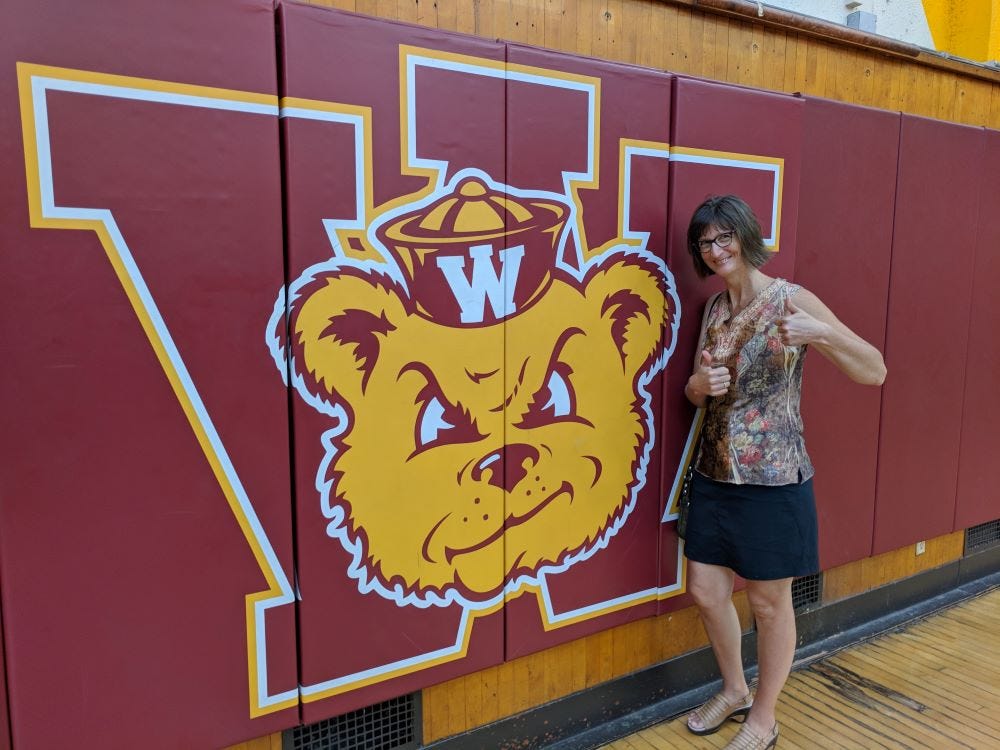 a middle-aged woman (me) standing next to a mat on the wall of a gymnasium, featuring a grumpy teddy bear face on a big letter "W"