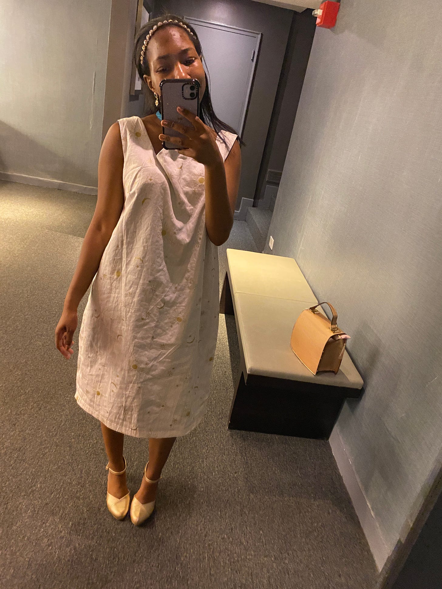 mirror selfie of a young woman wearing a white dress with gold moons and stars on it. The neckline is made of two v's. Behind her is her pink purse on a bench. She holds a lilac iphone with a transparent case