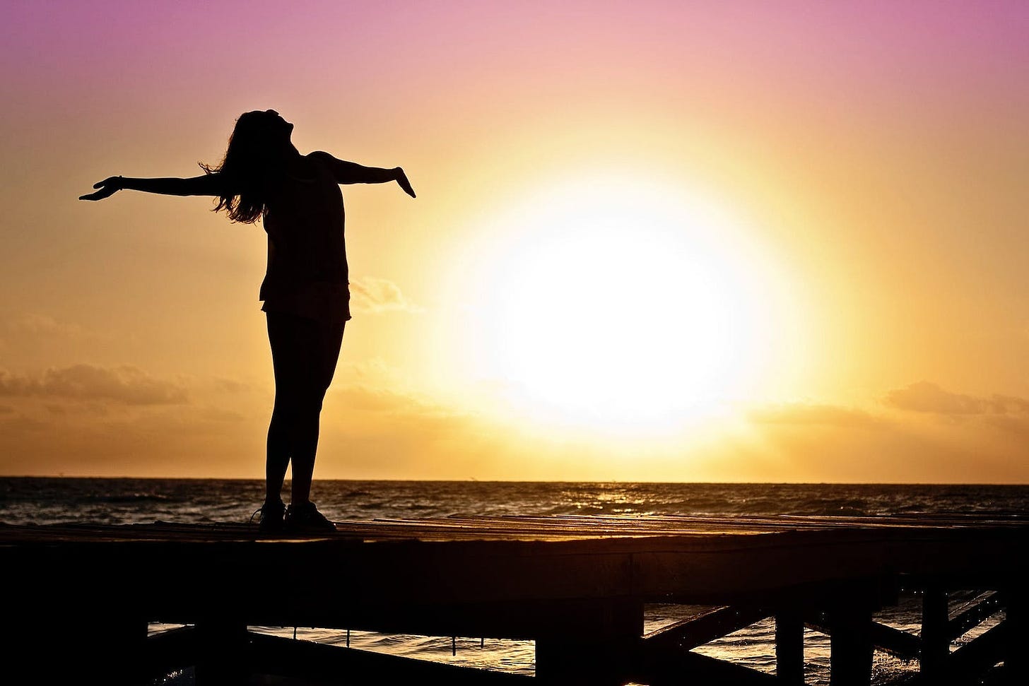Sillhouette of a young woman happy and with outstretched arms at sunrise or sunset. She has flowing long hair and with her head tilted back and eyes towards the sky.