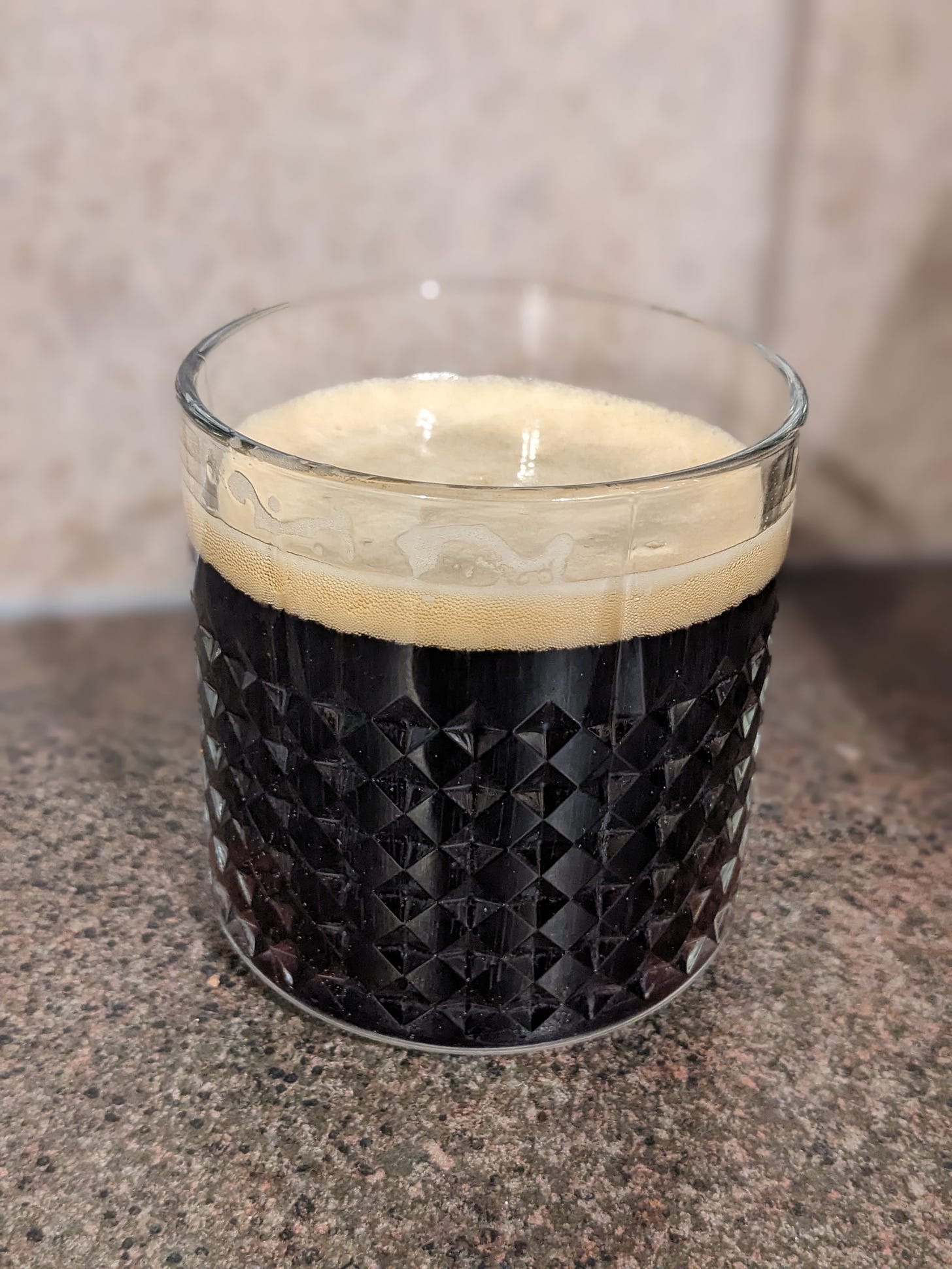 a glass of dark brown beer sits on a kitchen counter. the beer has light foam on top.