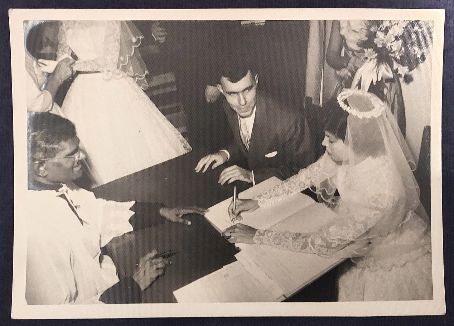 Black and white photo of a bride signing the register in front of a priest, and her young husband gazing at her