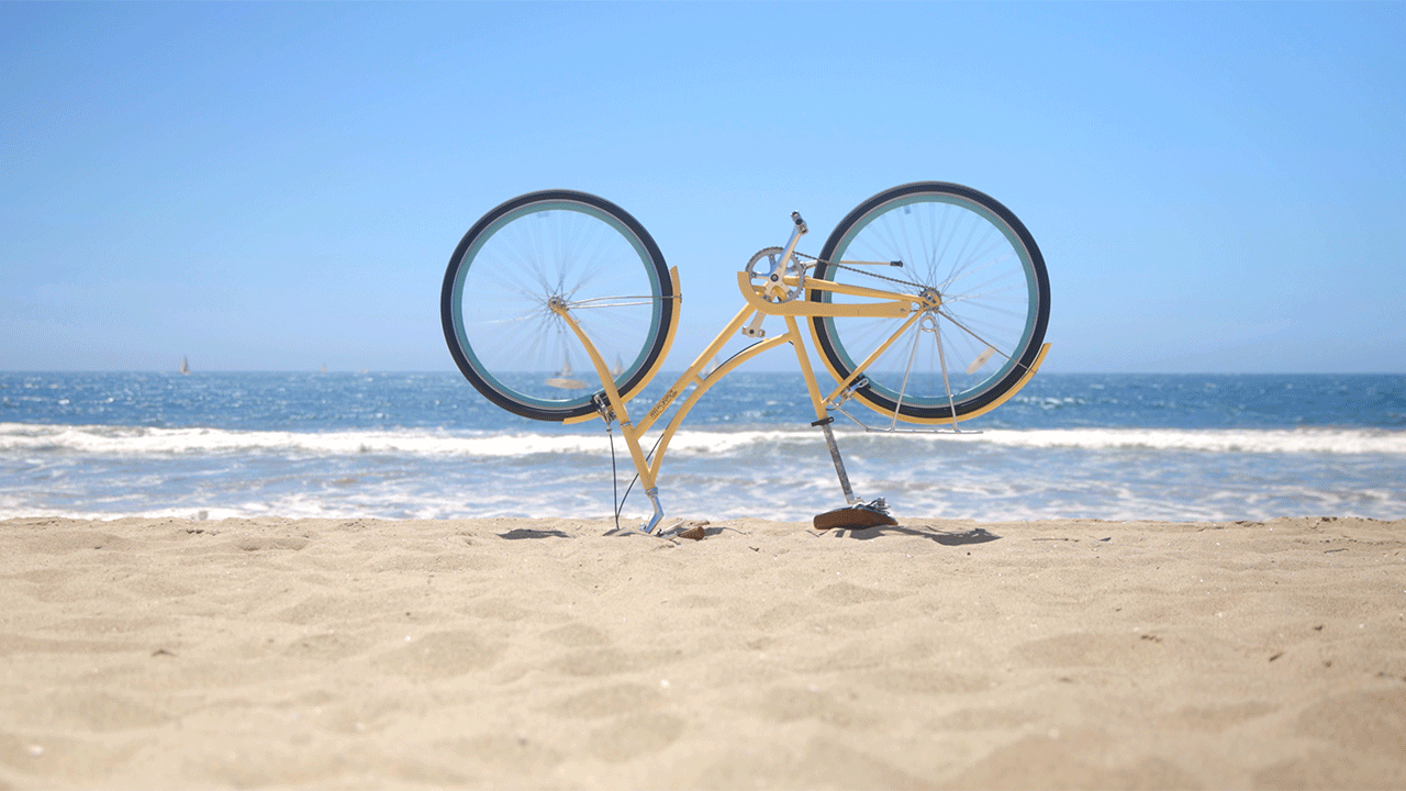 Gif de uma bicicleta de cabeça para baixo nas areias da praia, o mar ao fundo. As rodas giram em um movimento contínuo