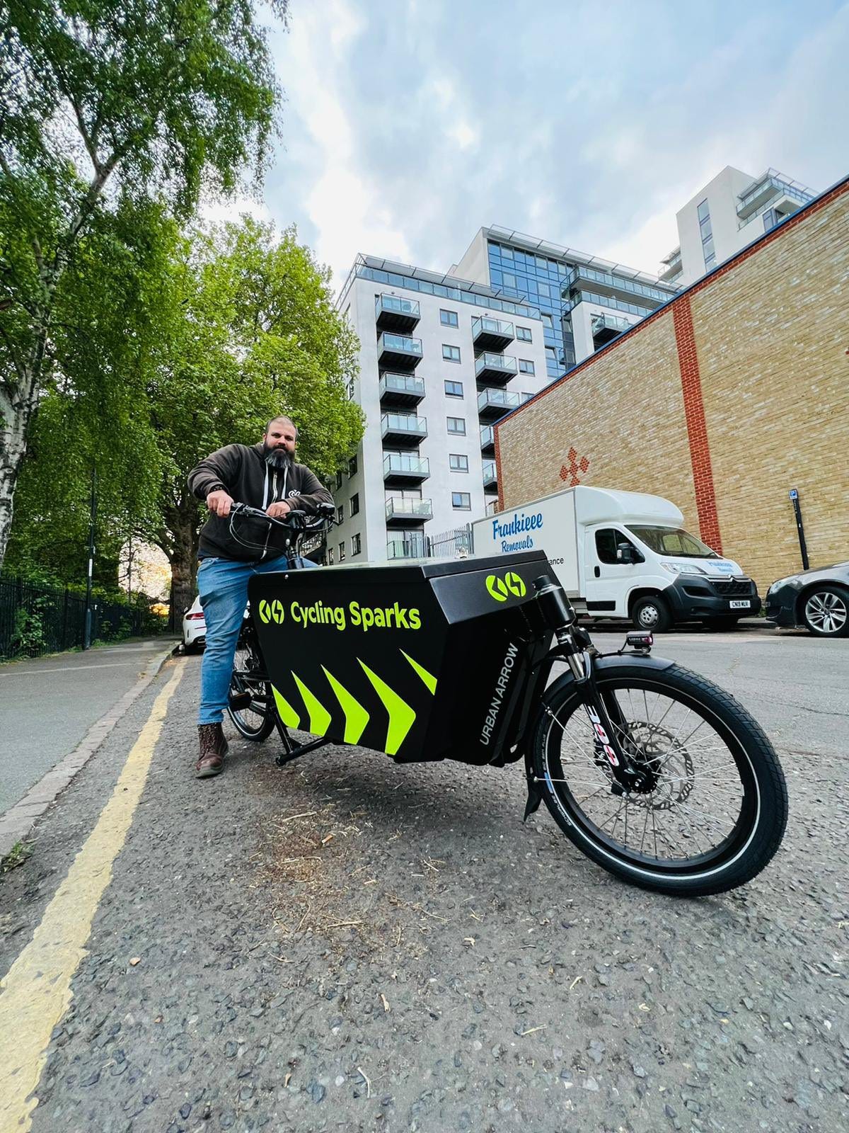 A man poses on the street whilst sitting on his cargobike. On the side of the cargobike is the name of his electrical company