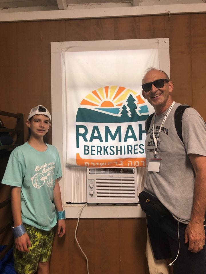 A camper wearing a baseball cap, tshirt and shorts stands with director Howard Blas who wears sunglasses, a tshirt and shorts