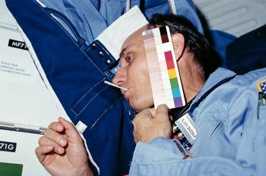 A pale man in a space shuttle holding up a palette of colors to his pale face.