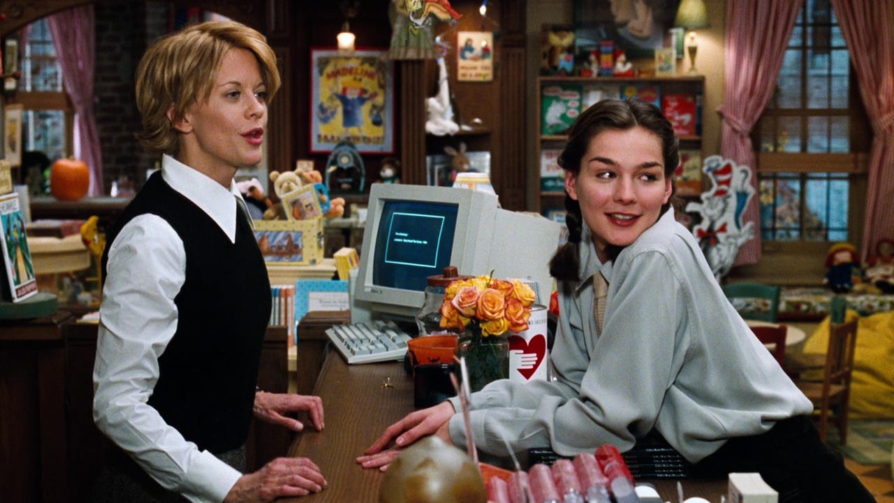 Still from the film you've Got Mail. Two white women stand on either side of a bookstore counter, looking at someone off-screen, orange flowers sit in a vase between them, an old 90's computer in the background.