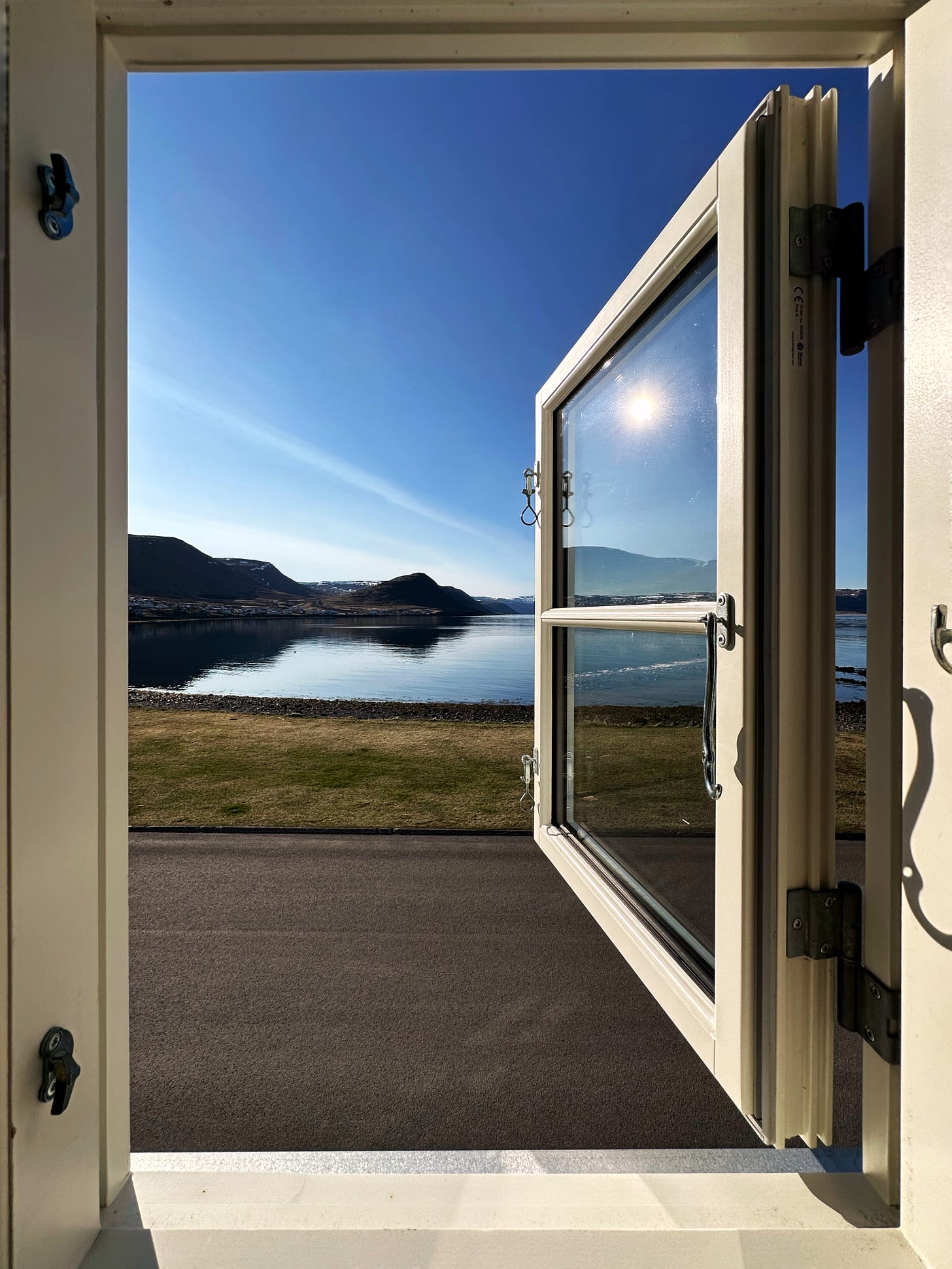 Morning sunshine through an open window overlooking a fjord in Patreksfjordur in West Iceland.  The sky is blue, the water is calm and the grass in the foreground is spring green.