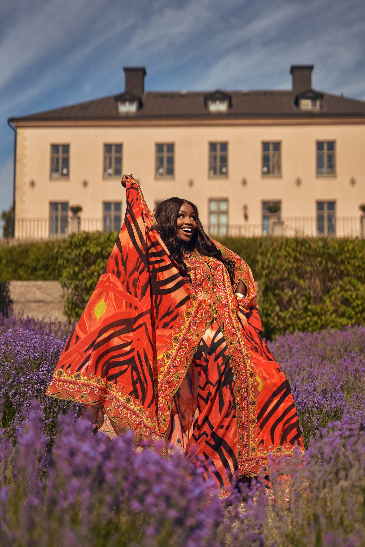 Lovette Jallow in bold patterned orange kaftan stands in a lavender field with arms raised, smiling in sunlight, exuding peace and autonomy.