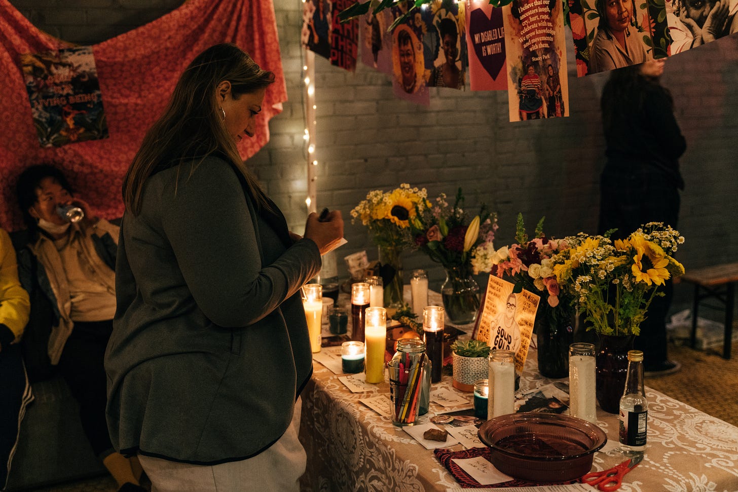 a person with olive skin and long dark hair looks down at the altar, clothespinned artwork images of disabled dead loved ones illuminated by tiny copper wire fairy lights above them, it's all candlelit.