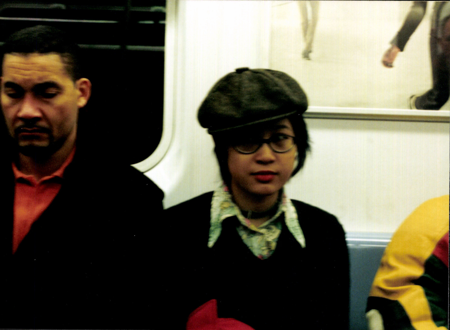 asian girl wearing a pageboy hat sirtting on the subway between two people, a man and an arm