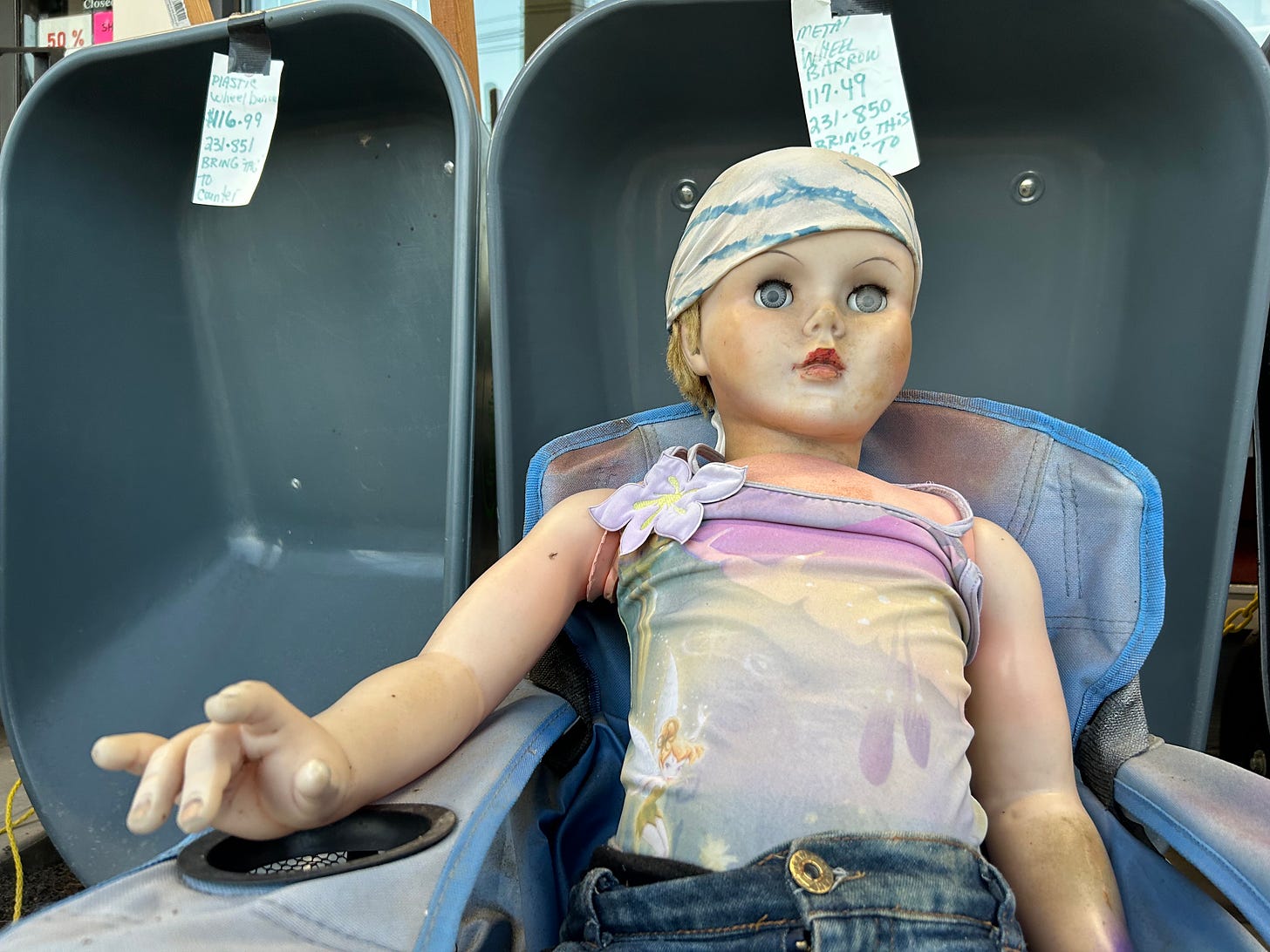 lifesize girl doll sitting in lawnchair in front of two wheelbarrows, wearing headscarf, tanktop, and jeans
