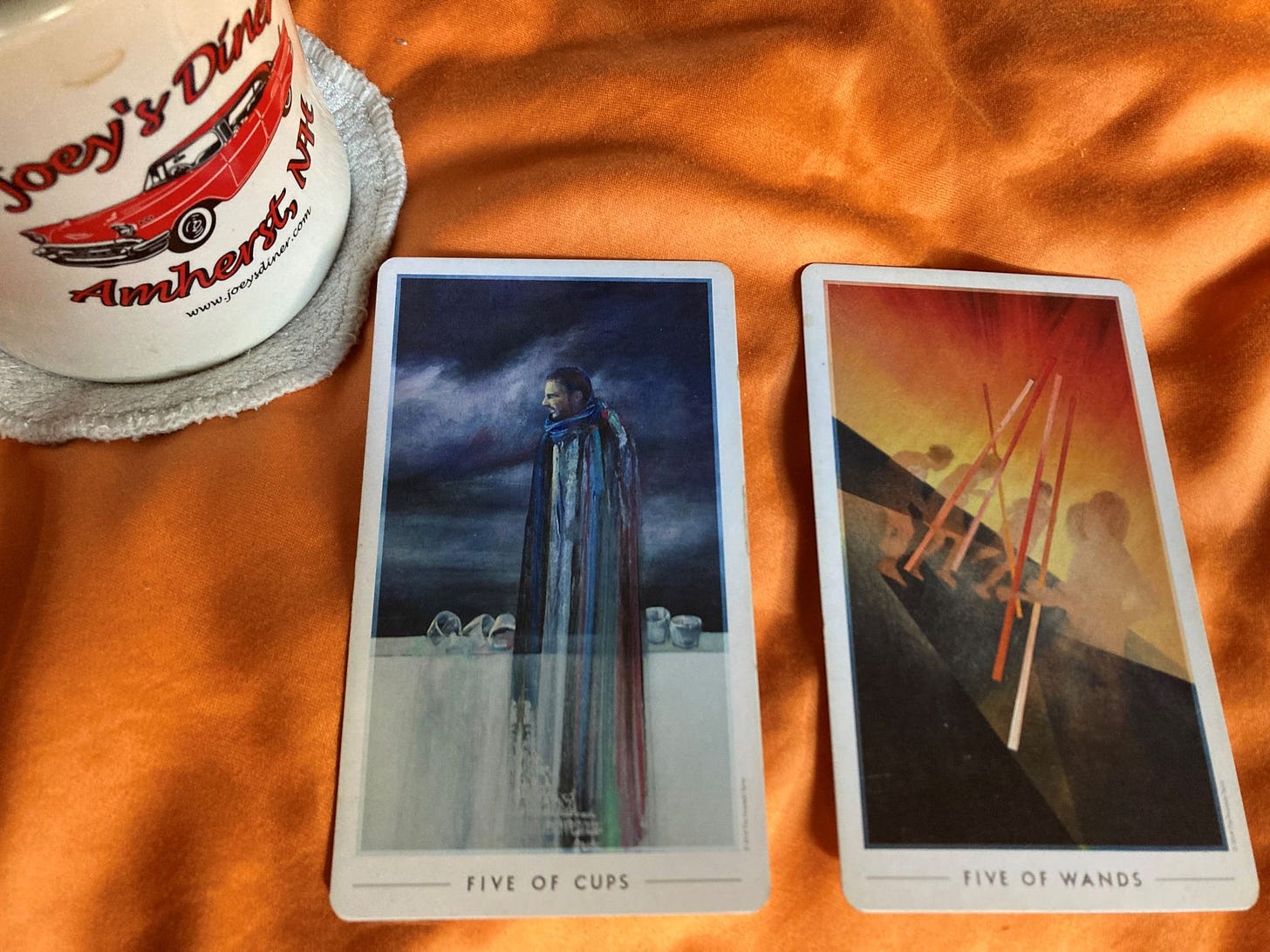 Five of Cups and Five of Wands Cards lie on an orange background with an empty coffee cup that sits on a used breast pad (which sums up my morning)