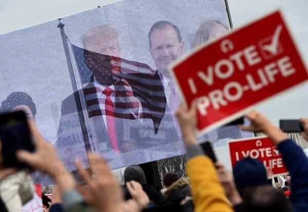 Demonstrators holding pro-life signs watch a large outdoor screen showing Donald Trump speaking to the crowd. The screen is fading between a shot of Trump and a shot of the American flag; both are visible, layered over each other.