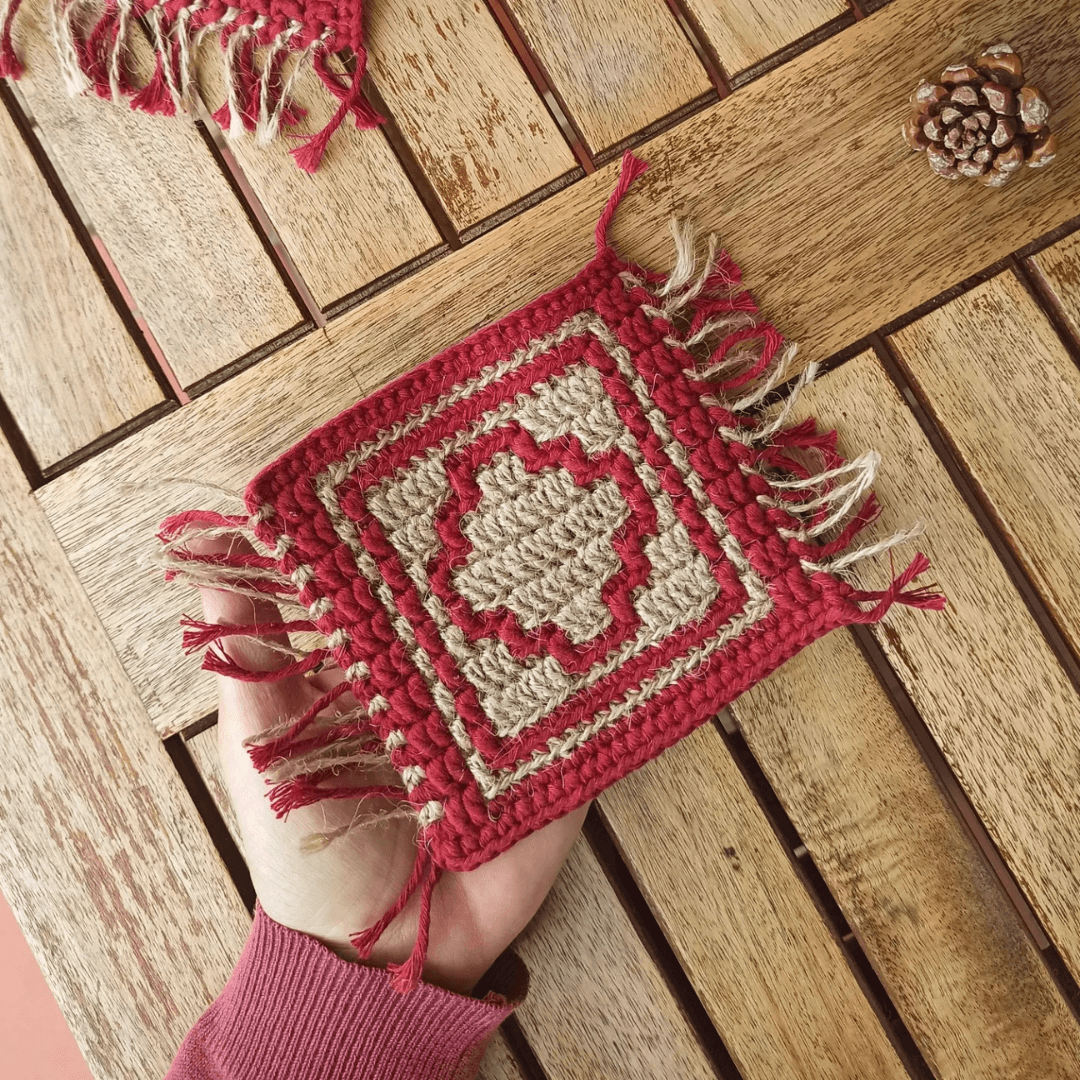 Square coaster in dark red cotton and jute made in mosaic crochet with a geometric design and fringes. Hand is holding it over a wooden table top.