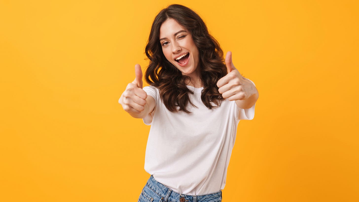 Smiling woman in a white t-shirt giving two thumbs up against a bright orange background.