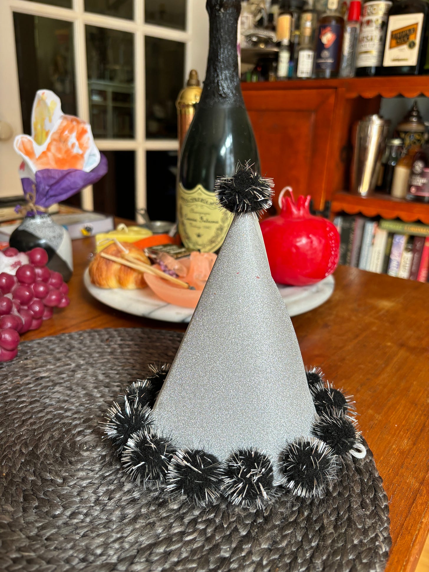 Table with a gray festive party hat set atop a gray placemat. Champagne and candles in the background