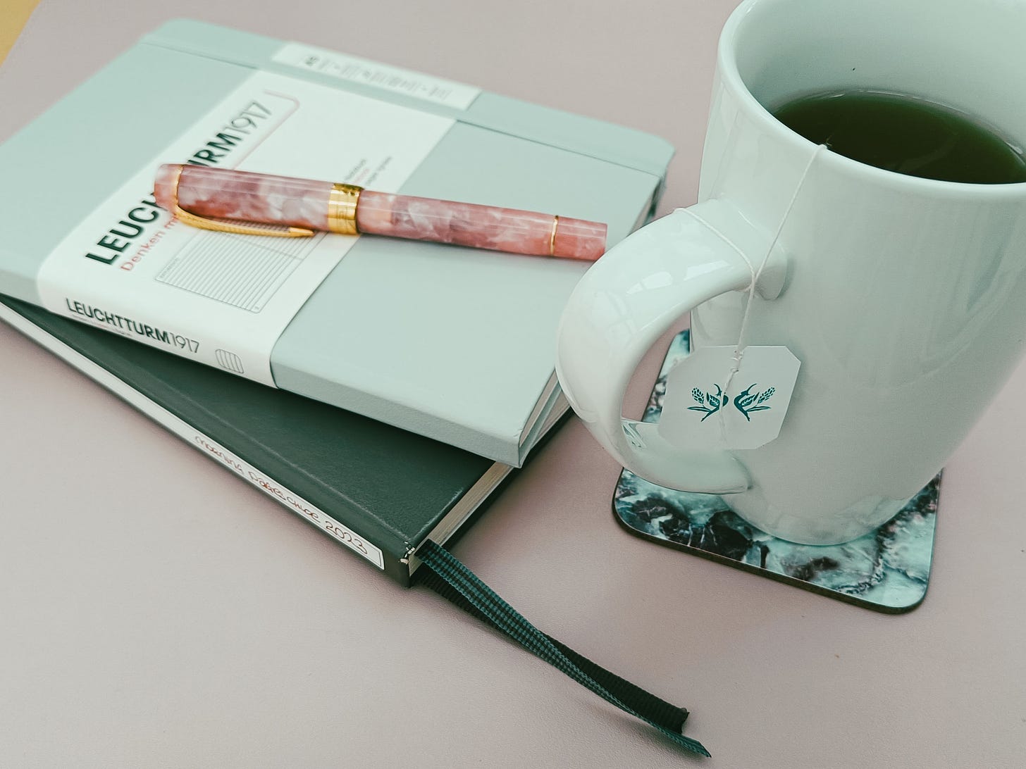 Img: Stack of 2 A5 Leuchtturm1917 journals with a pink and gold fountain pen on top, plus a tall white mug of herbal tea.