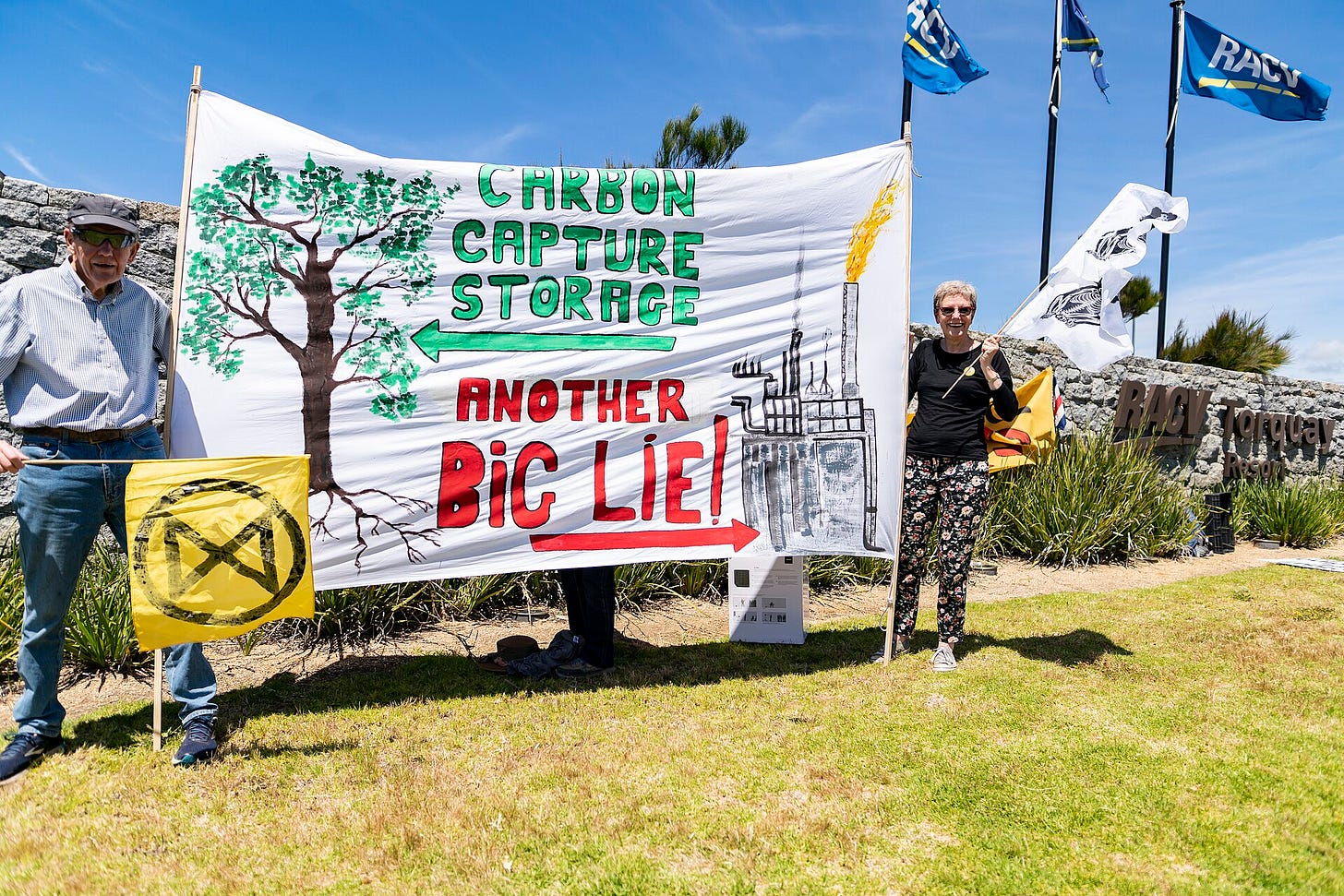 Cloth banner being held up by two people. The banner has a picture of a tree with an arrow pointing to it saying "Carbon capture storage". The banner also has a picture of an industrial facility and an arrow pointing to it saying "Another big lie".