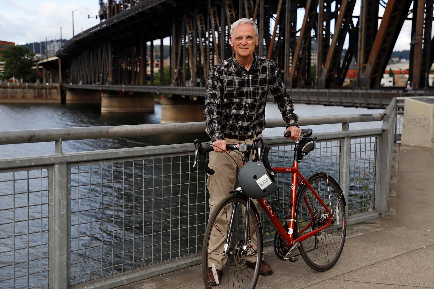 The Congressman standing with his red bike by a river and a bridge.