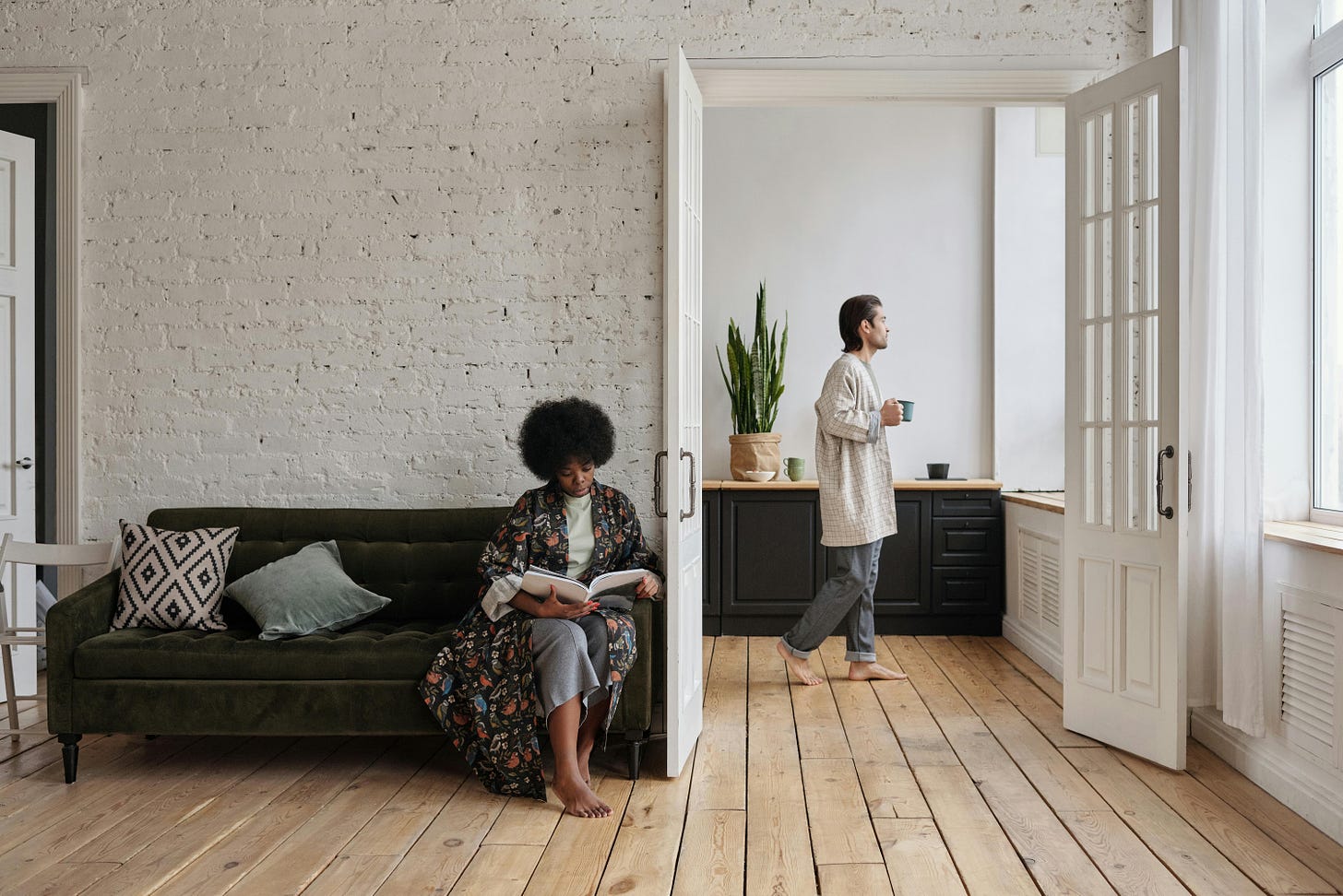 Two rooms with a white brick wall and open two-door doorway between them. A Black woman with Afro sits on a black couch reading, and in the room behind seen through the open doorway, a White man walks with blue coffee cup towards a window. Neither faces towards each other.