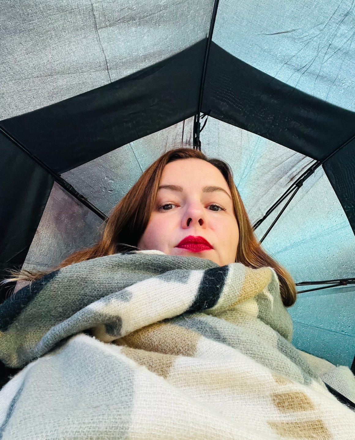 Amber takes a selfie. The phone is low, pointing upward at her face. She has an open umbrella over her head, covering the background of the photo. She wears a scarf and red lipstick. 