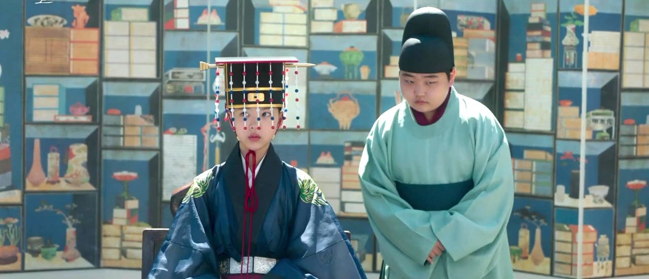 A young girl dressed as a crown prince and a young eunuch in front of a screen painted with books