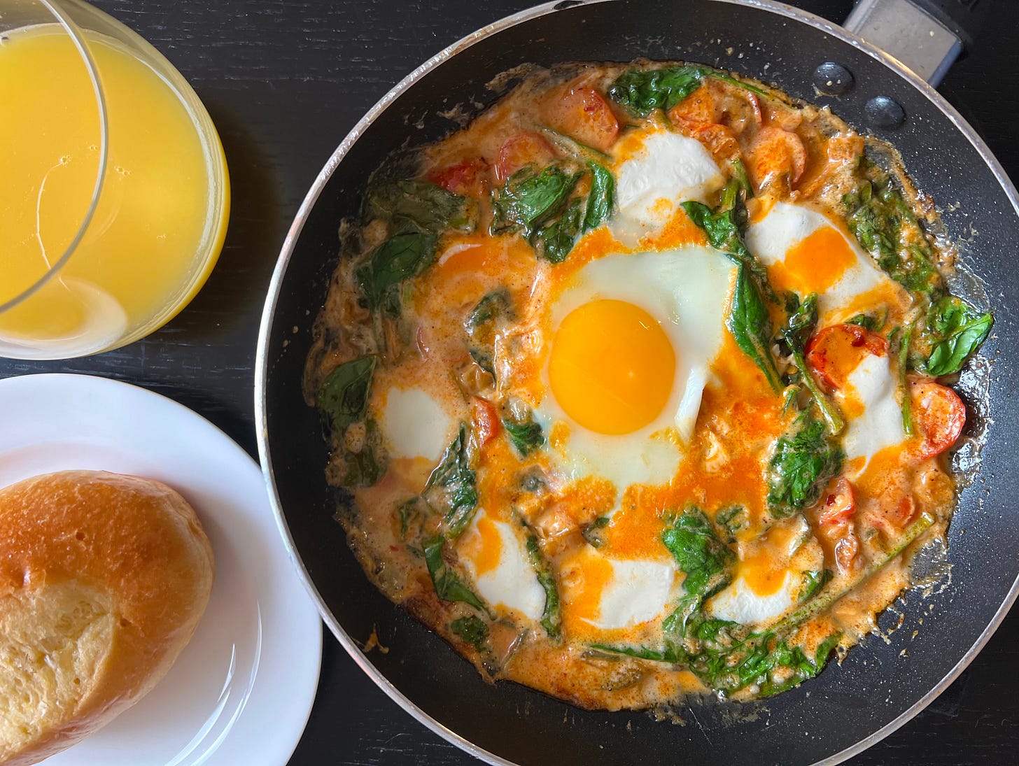A freshly made shakshuka with vibrant red tomatoes, wilted spinach, and melted mozzarella, topped with a bright golden egg yolk. The dish is served with a soft roll and a glass of fresh orange juice, making for a perfect breakfast or brunch. Liat Portal made this as part of her Foodie Disorder obsession, experimenting with different shakshuka variations.