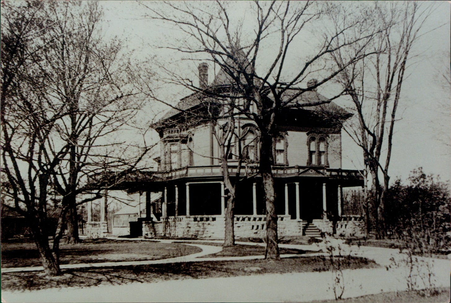 Black and white photo of Victorian house with wrap-around porch and steeply pitched hip roof with corner tower topped by a pointed roof.