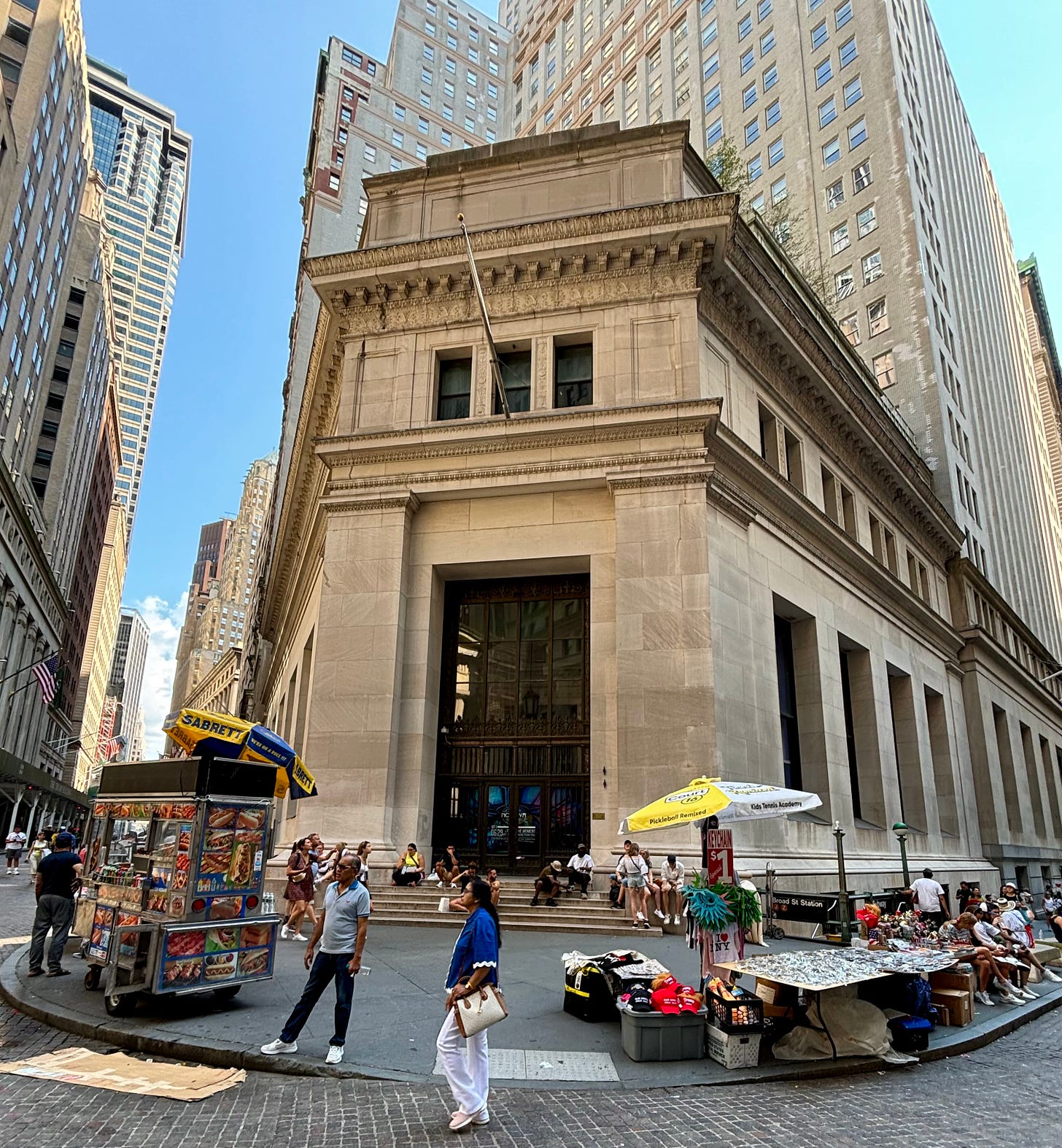 A low, neo-classical building on a corner surrounded by large office buildings. Tourists lounge on the steps, and there are hot dog and pretzel carts and a seller with a table of cheap junk including MAGA hats with sewn in wigs, all waiting for the tourists to bite.